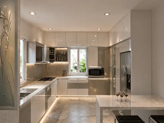 L-shaped modular kitchen in glossy white with a built-in microwave, a steel refrigerator and a white breakfast counter in the foreground